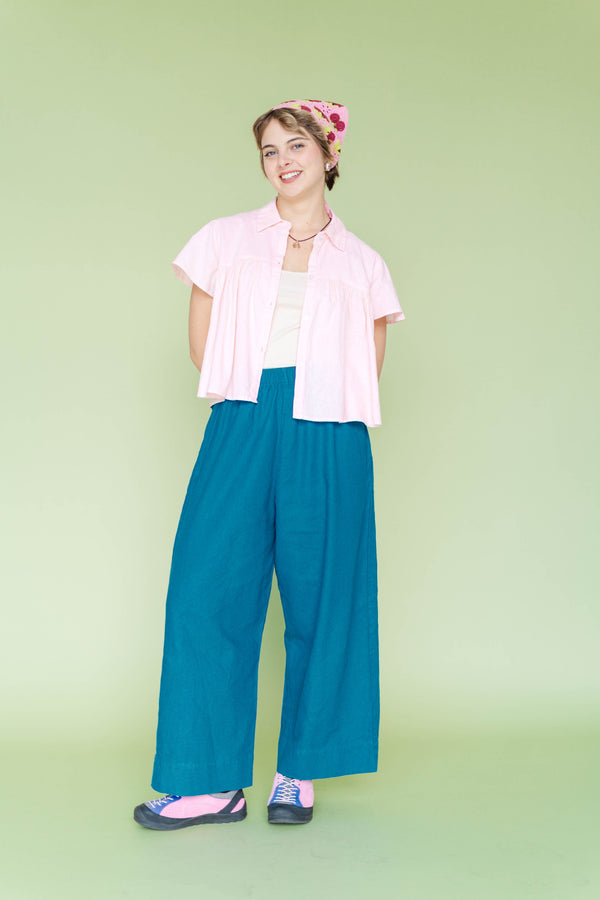 Full-length studio view of a model wearing Nettle Studios Teal Mid Easy Pants paired with a light pink short-sleeve button-up top and sneakers.