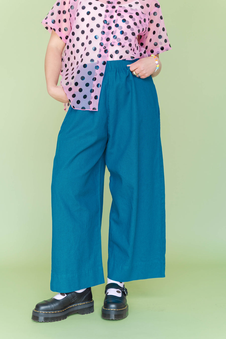 Mid-section close-up highlighting the elastic waist, side pockets, and wide-leg cut of the teal linen pants styled with a sheer polka-dot shirt.