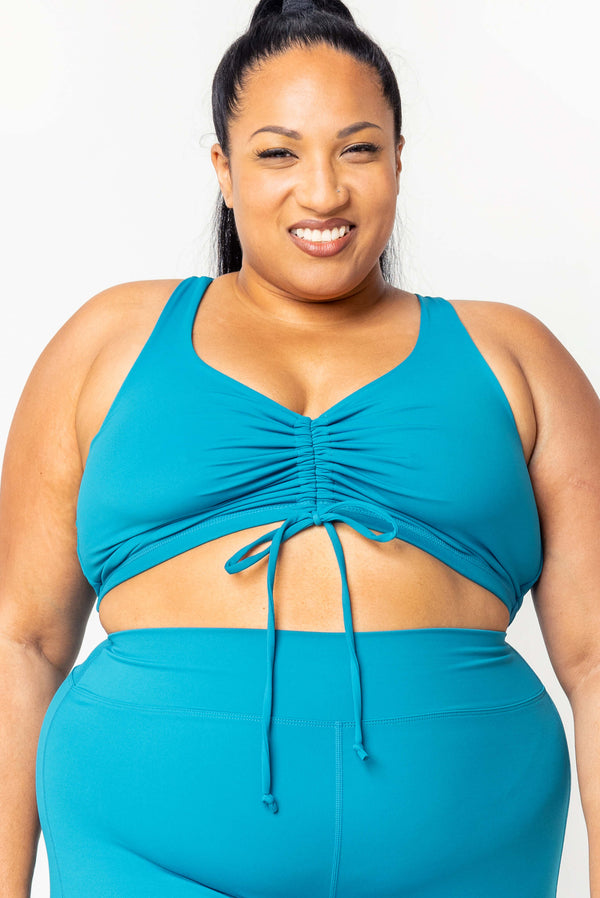 Front view close-up of a smiling plus-size model wearing the teal Superfit Hero Cinched Bikini Swim Top with front cinch tie paired with high-waisted teal swim bottoms.