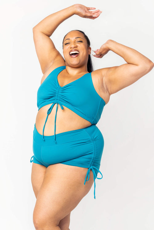 Three-quarter full-body view of a model laughing with arms raised, wearing the teal cinched bikini top and matching cinched swim shorts.