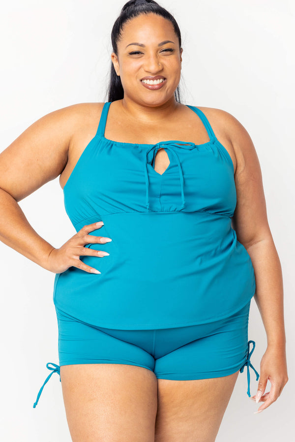 Front view of a smiling plus-size model wearing the teal Superfit Hero Tankini Swim Top with front cinch tie and matching teal swim shorts.
