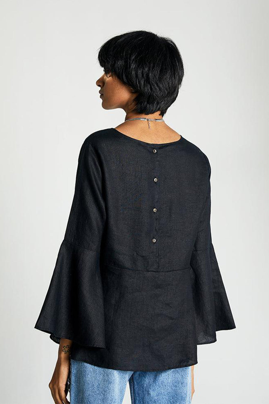 Back profile view of a straight-size model displaying the delicate row of buttons along the spine of the black hemp top, showcasing the elegant proportions of the flared sleeves from behind.