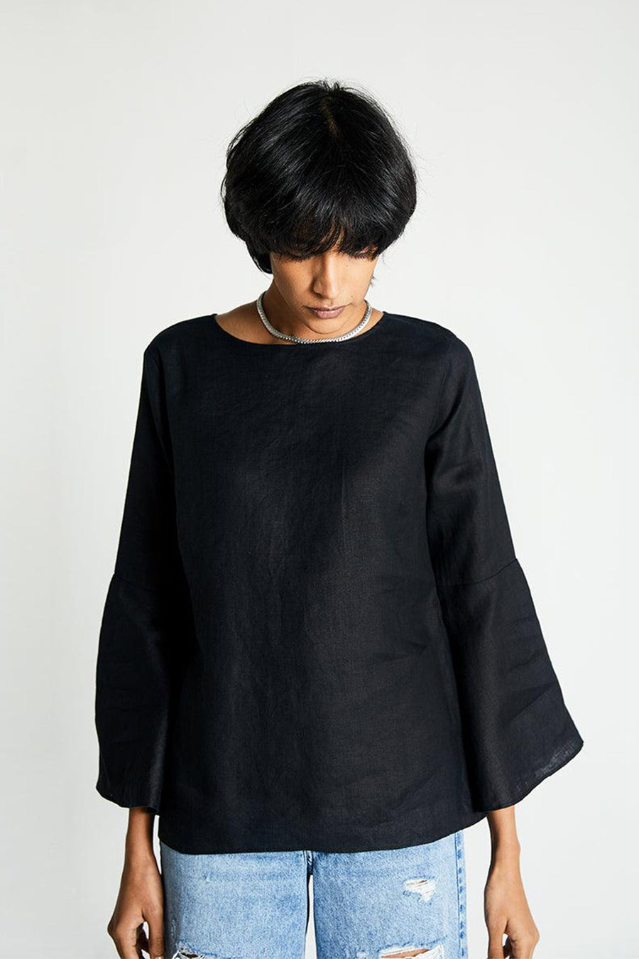 Front mid-shot of a model looking down, highlighting the neat lines of the boat neck design and the understated everyday elegance of the black Reistor linen-look shirt.