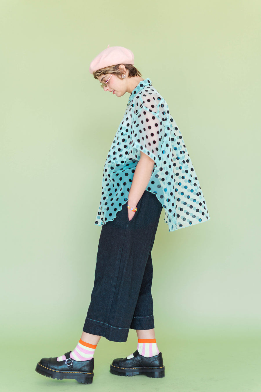 Side profile view of a model walking, showcasing the dramatic flared drape and unique curved petal silhouette of the Nettle Studios Sheer Mint Chocolate Chip Polka Dot Tulip Top in a size-inclusive fit.
