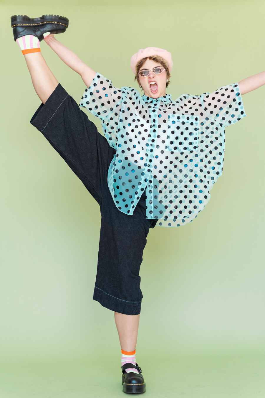 Model performing a high leg extension wearing the Nettle Studios Black Daisy Denim wide-leg pant, styled with a light blue sheer polka dot blouse and beret.