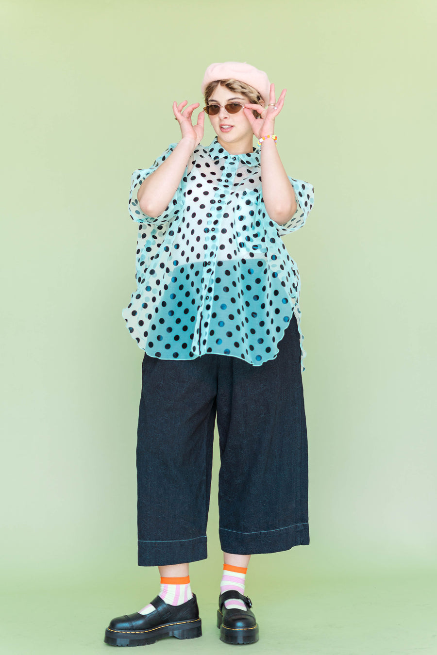 Full-body view of a model in a size-inclusive fit wearing the Nettle Studios Sheer Mint Chocolate Chip Polka Dot Tulip Top. The short-sleeve button-up is styled over dark blue cropped trousers against a solid lime green background, showcasing its sheer minty organza volume.