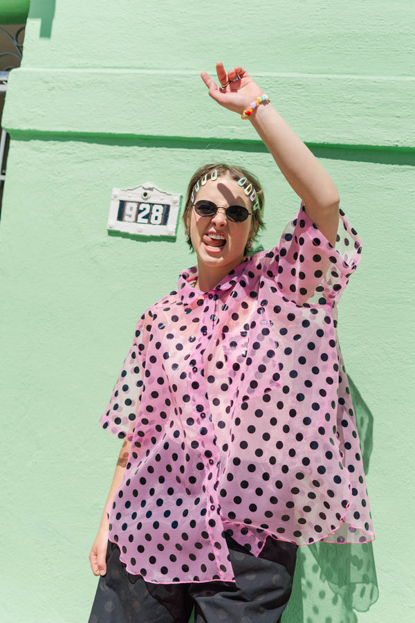 Model outdoors wearing the pink sheer polka dot tulip top by Nettle Studios, styled with dark trousers and oval sunglasses.