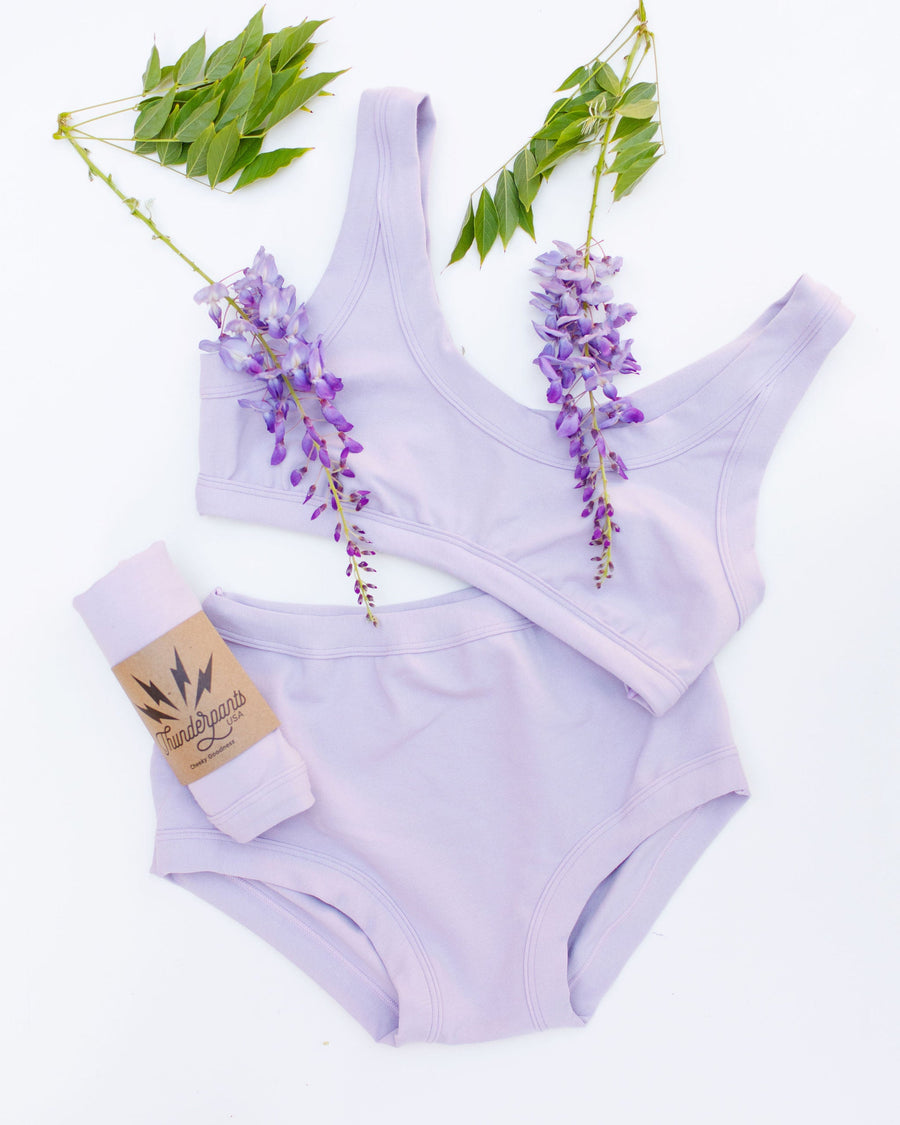 Flat lay product display showing the Wisterial Girl bralette, full briefs, and rolled Thunderpants US packaged underwear with fresh wisteria blooms.