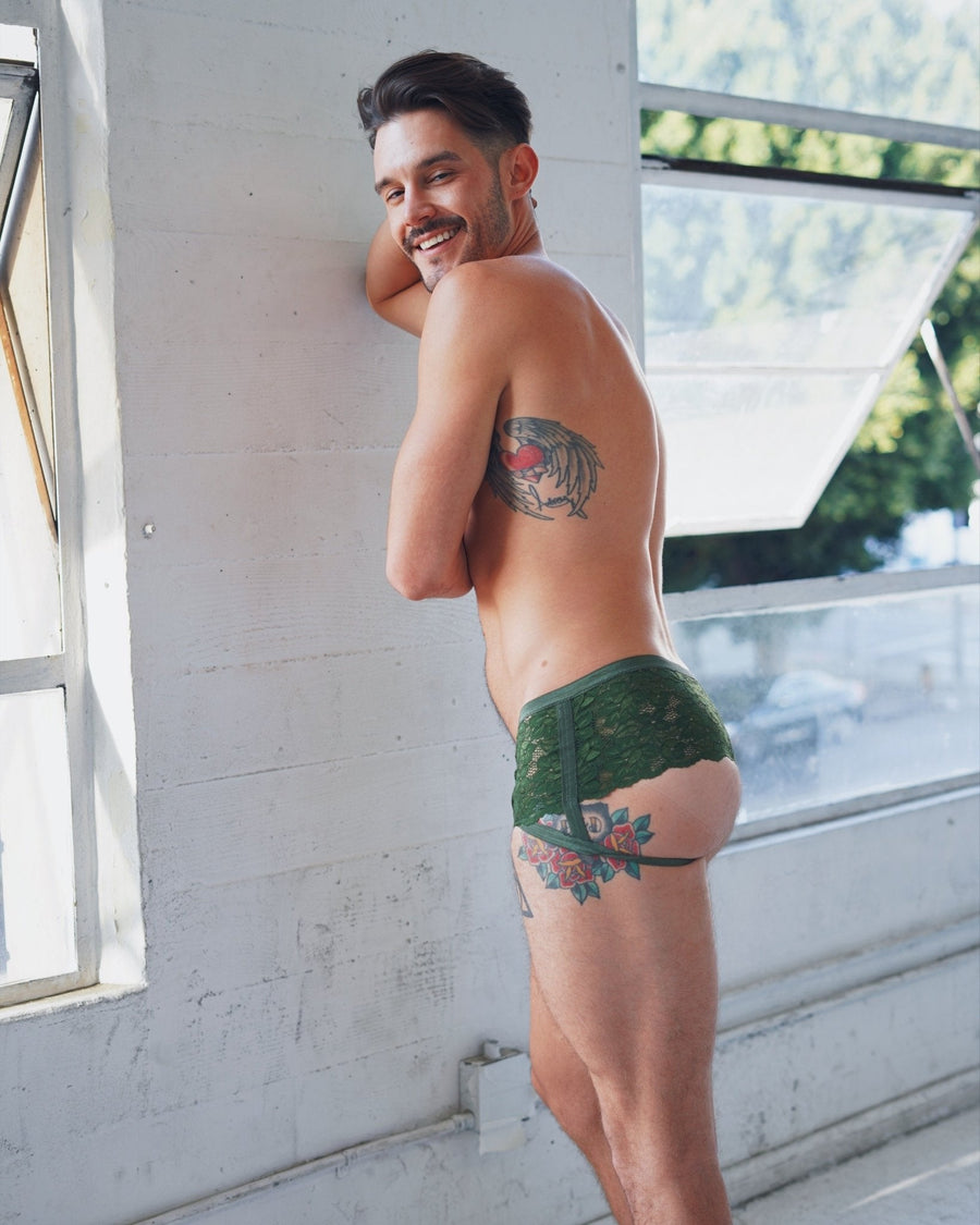 Side view profile of a brunette male model wearing dark green lace cantiqLA Bandage Boyshorts leaning against a white studio wall.
