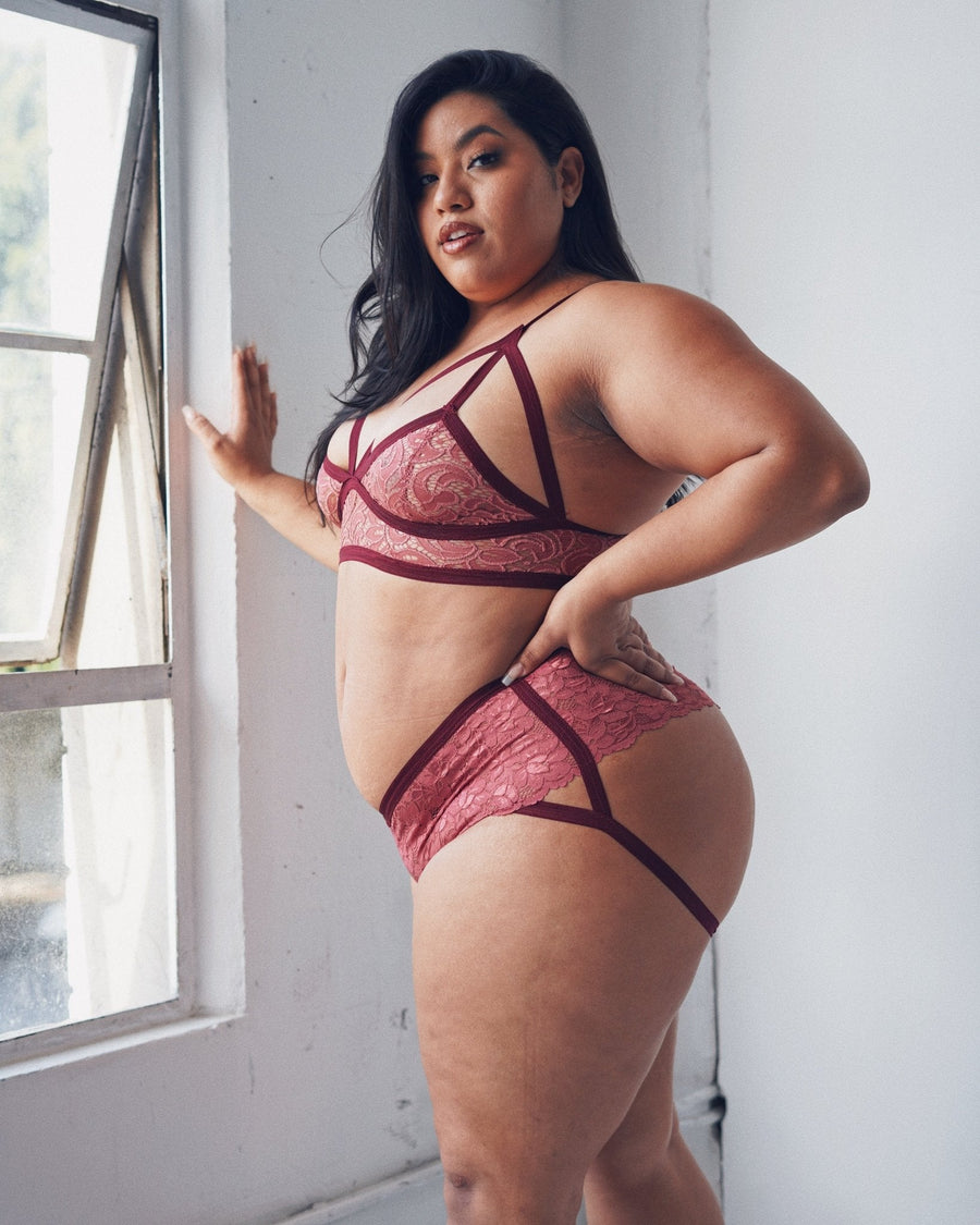 Side profile portrait of a model standing by a window wearing plum-colored lace cantiqLA Bandage Boyshorts.