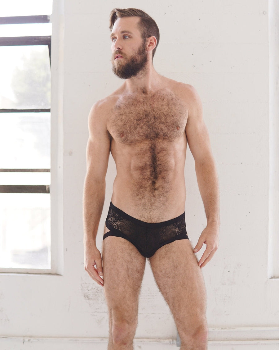 Front view of a bearded male model wearing the size-inclusive black lace cantiqLA Bandage Briefs against a white studio wall.