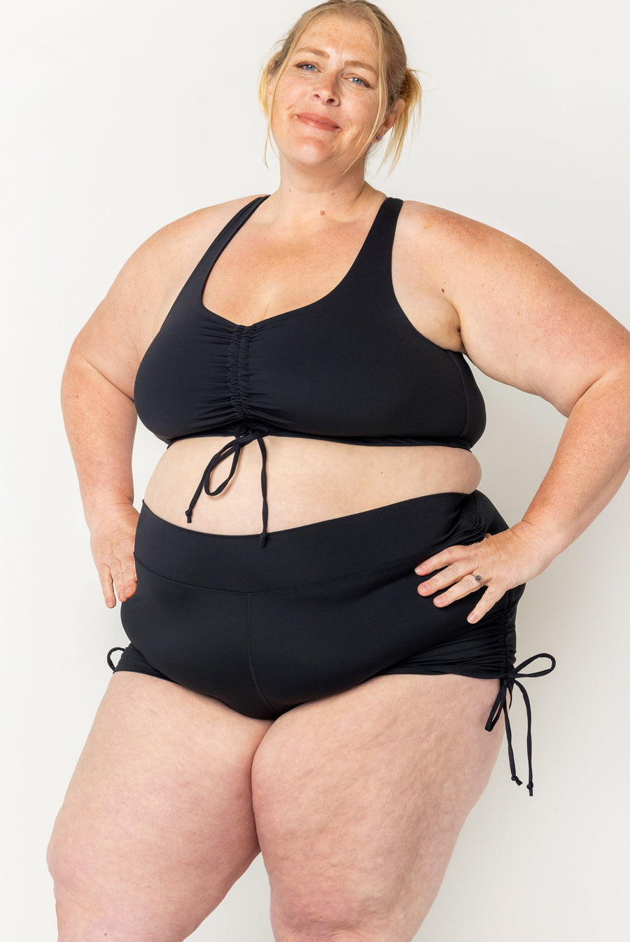 Front view of a blonde plus-size model posing with hands on hips wearing black high-waisted swim booty shorts with side cinch ties.