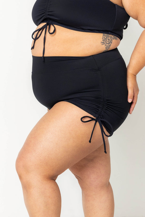 Side view close-up of a plus-size model showing the cinched side tie detail on high-waisted black Superfit Hero swim booty shorts.
