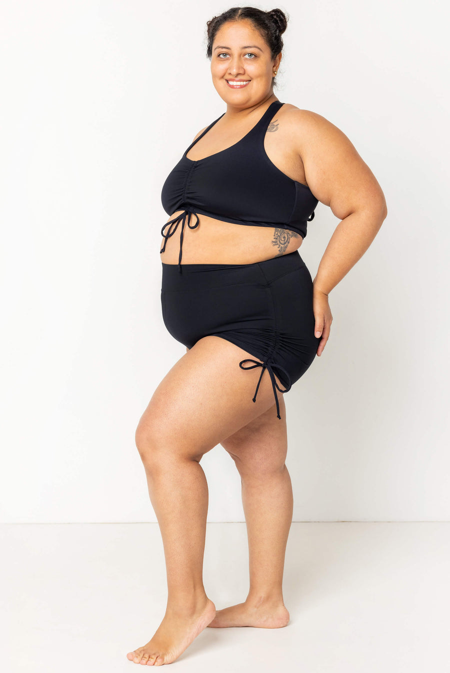 Full-length side profile of a model posing in high-waisted black swim booty shorts with side ties paired with a matching black swim top.