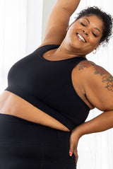 Side view of a smiling plus-size model with shoulder tattoos stretching upwards in the black full-coverage SuperHold Racerback Sports Bra.