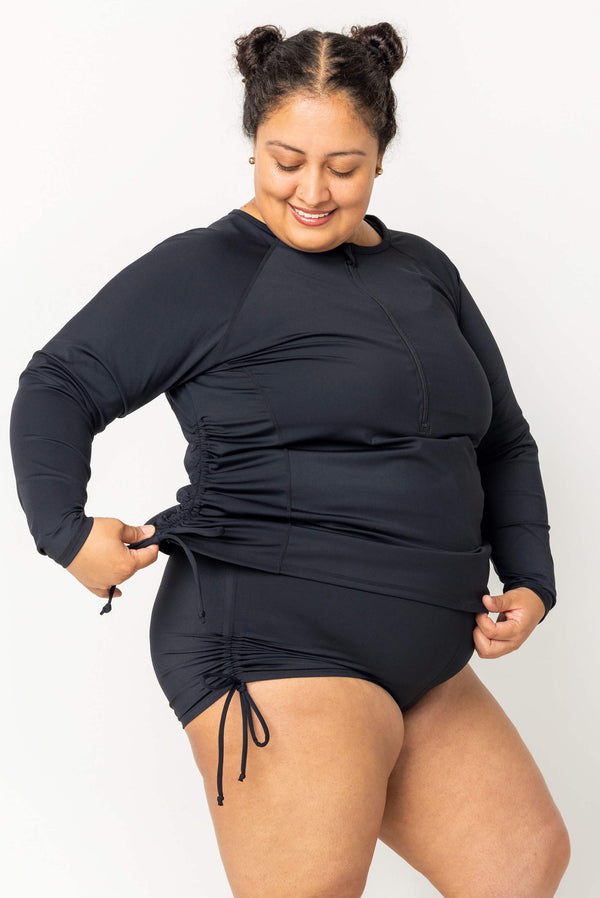 Side-front view of a smiling plus-size model adjusting the adjustable side cinch tie on the black Superfit Hero long sleeve swim rash guard.