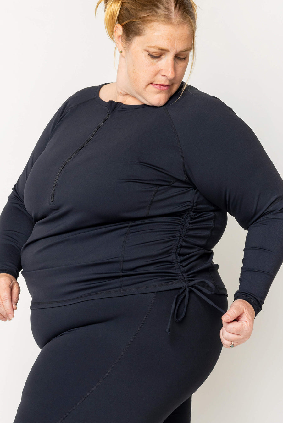 Three-quarter side shot of a blonde model showing the cinched side-ruched drawstring detail on the black long sleeve swim top.