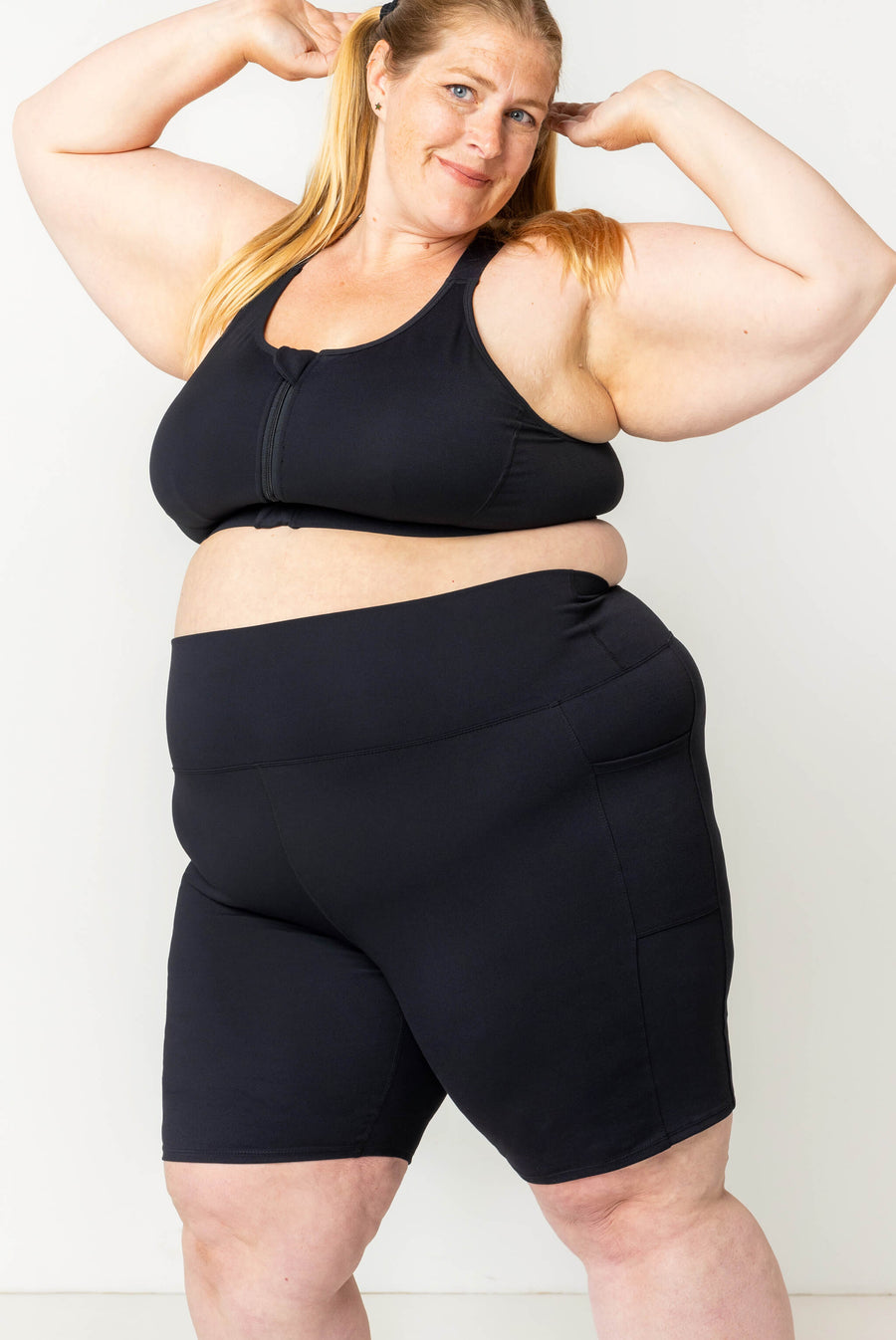 Three-quarter shot of a blonde model flexing her arms while wearing high-waisted black 9-inch compression shorts and a matching black zip-front sports bra.