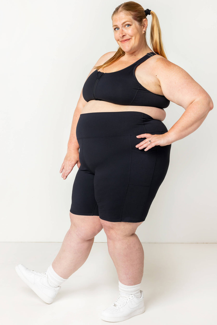 Full-body side profile of a blonde model wearing black Superfit Hero SuperHold 9-inch shorts, a black sports bra, and white sneakers.