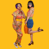 Full-length studio view of a model in the Blue Platypus Brownstone Betties Jumpsuit paired with white sandals against a bright yellow wall.