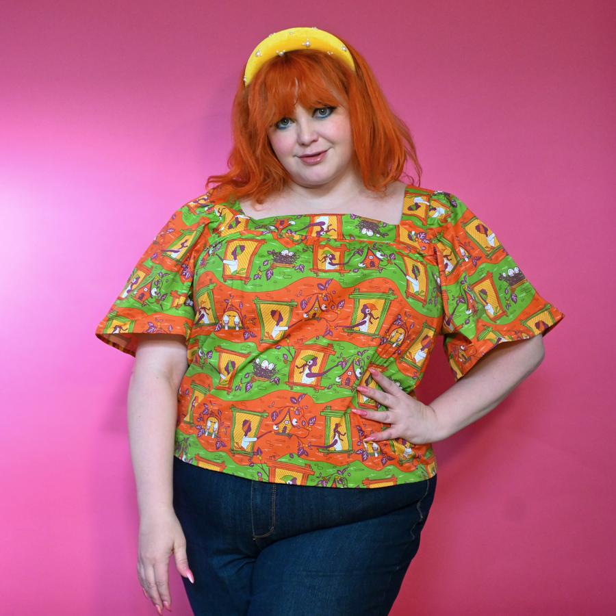 Model wearing the Pumpkin & Olive Brownstone Betties top holding a fluffy cat against a pink studio background.