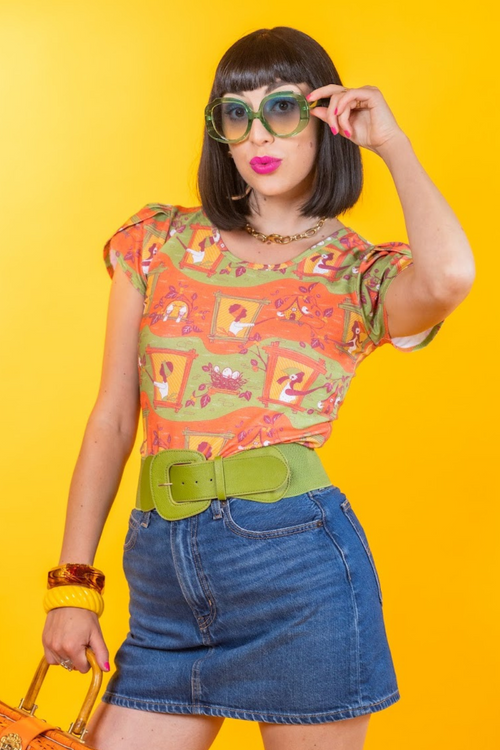Studio portrait of a dark-haired model with green sunglasses wearing the Blue Platypus Brownstone Betties Tulip Sleeve Tee in Pumpkin & Olive against a yellow backdrop.