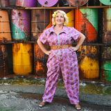 Model posing outdoors against colorful background barrels wearing the size-inclusive Blue Platypus Cheeky Tiki Monkeys Jumpsuit in Magenta.