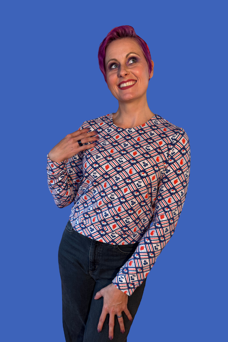 Studio portrait of a model with short purple hair wearing the Blue Platypus Fruit Picnic Pullover in Blueberry against a blue backdrop.