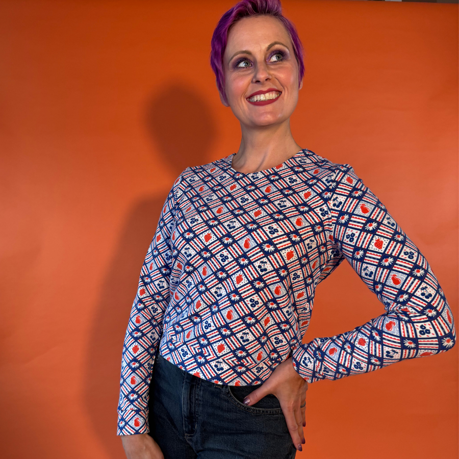 Model with purple hair posing in dark jeans and the Blue Platypus Fruit Picnic Pullover in Blueberry against an orange studio background.
