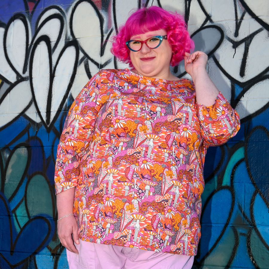 Model with pink curly hair posing in front of a graffiti heart wall wearing the Blue Platypus Groovy Mushroom 3/4 Sleeve Tee in Sunset.