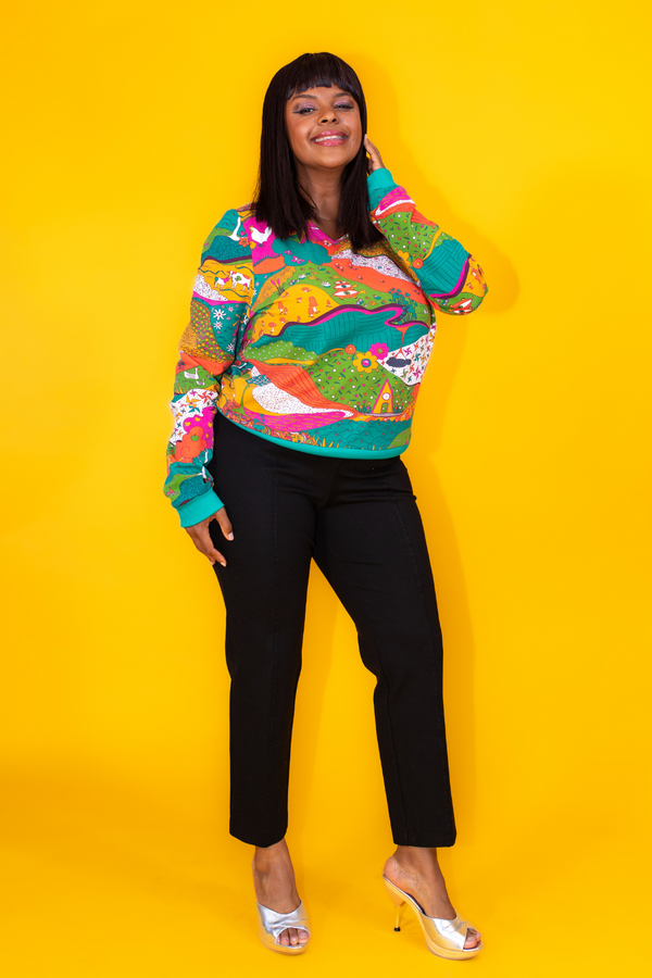 Full-length view of a model in the cropped Blue Platypus Landscape Pullover in Emerald styled with black trousers and silver heels.