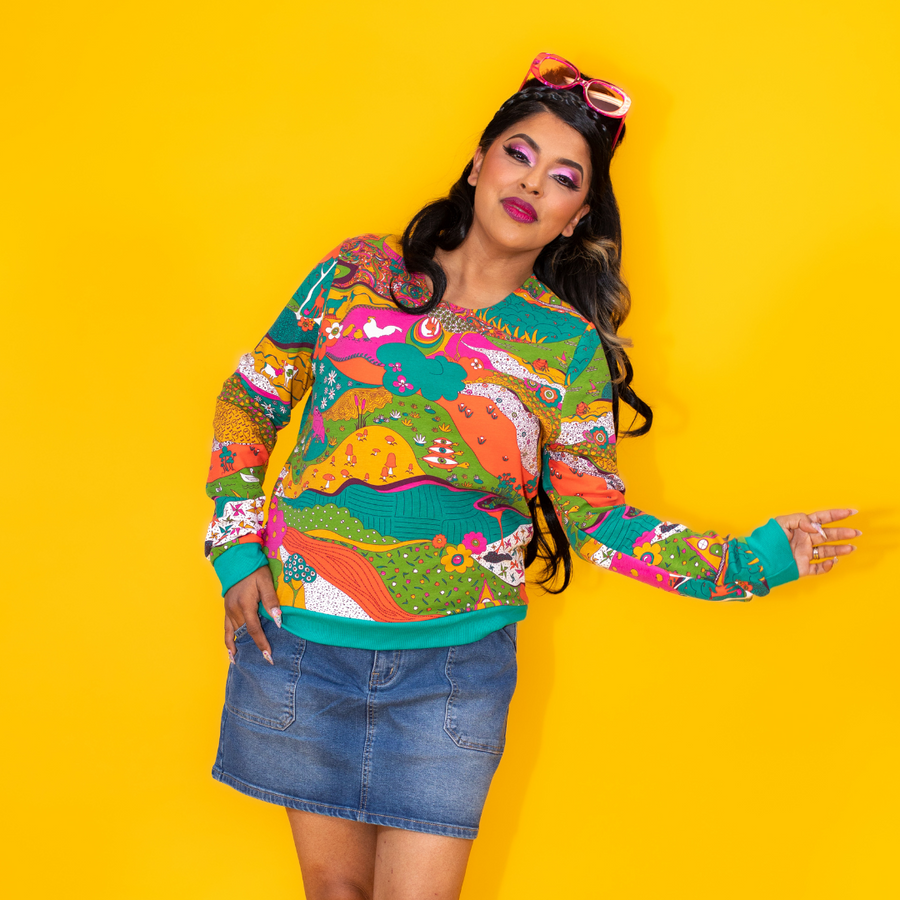 Studio portrait of a model wearing the Blue Platypus Landscape Pullover in Emerald paired with a denim mini skirt against a yellow backdrop.