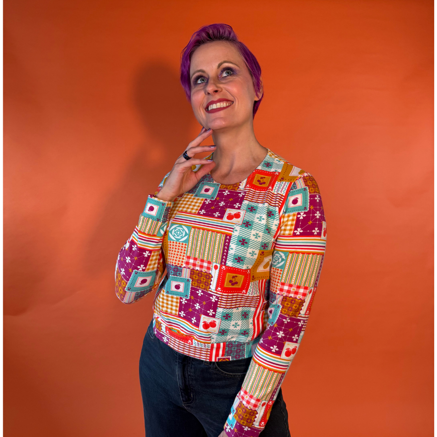 Model with purple hair posing thoughtfully while wearing the Blue Platypus Patchwork Pullover in Magenta against an orange backdrop.