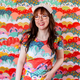 Studio portrait of a smiling woman with glasses wearing the Blue Platypus Tulip Sleeve Top in Mint & Strawberry Meadow Print against a colorful background.