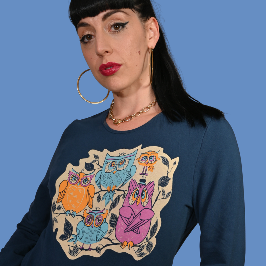 Studio portrait of a dark-haired model wearing the Blue Platypus Wacky Owls Pullover in Midnight Blue styled with a brown plaid skirt.