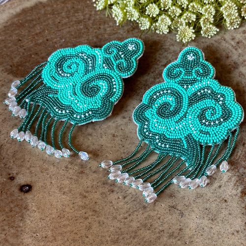 Studio view of the aqua beaded cloud earrings resting on a textured ceramic plate alongside dried white flowers.