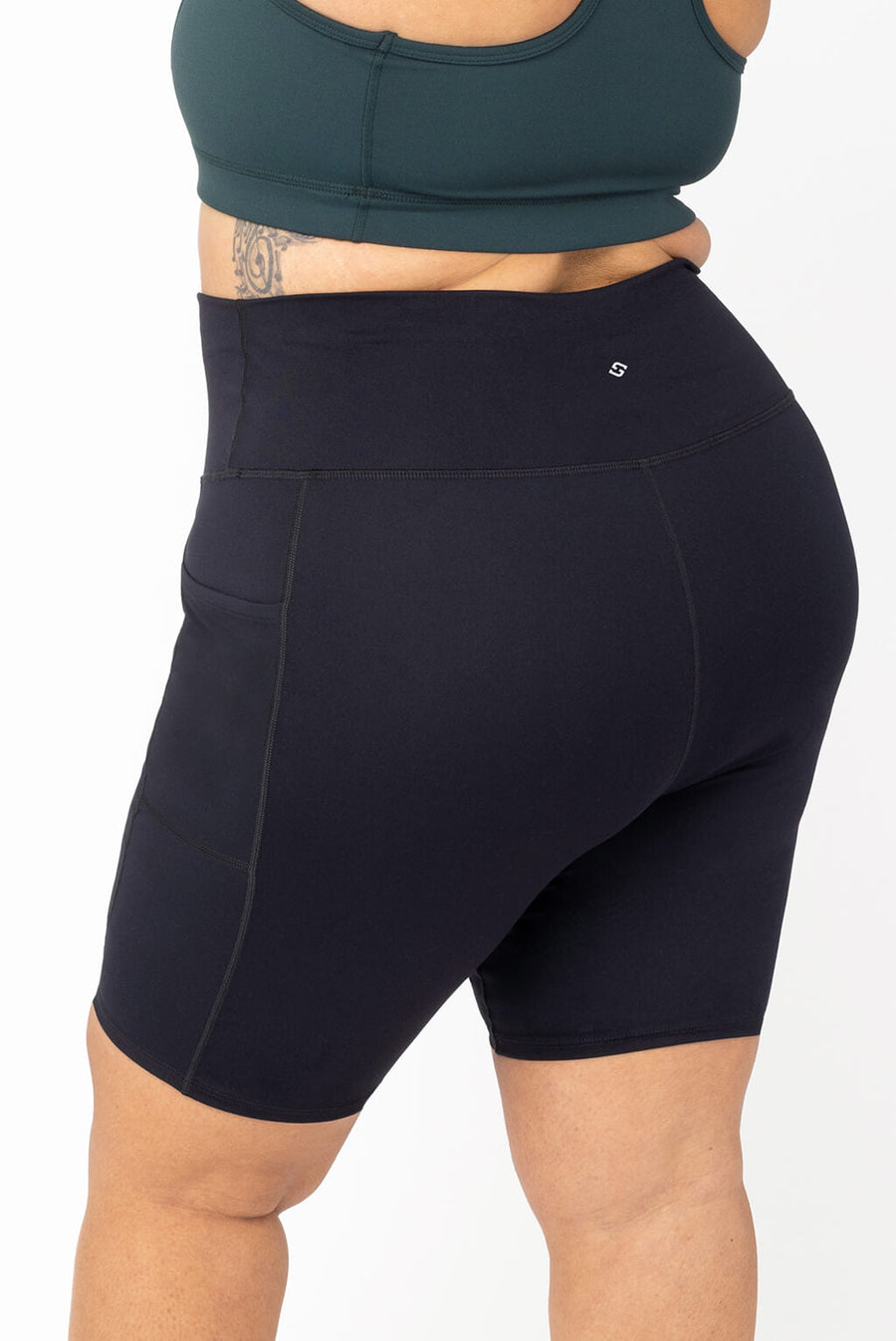 Side-back view of high-waisted black SuperHold 9-inch compression shorts with side pockets on a plus-size model wearing a dark green sports bra.