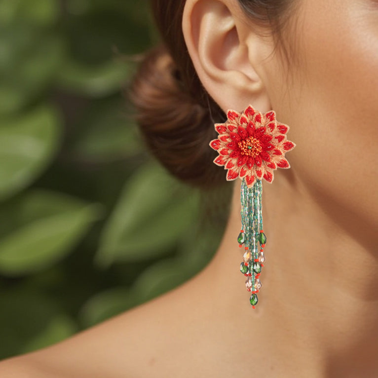 Close-up shot of a model wearing the Coral Red DAHLIA Embroidered Earring by Fyoli Fyoli. The image showcases the lightweight 3D flower design and green beaded fringe elegantly framing the jawline against a blurred green background.
