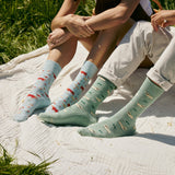 Two people sitting together outdoors on a picnic blanket, one wearing light blue farm animal socks and the other wearing green Valais Blacknose sheep socks.