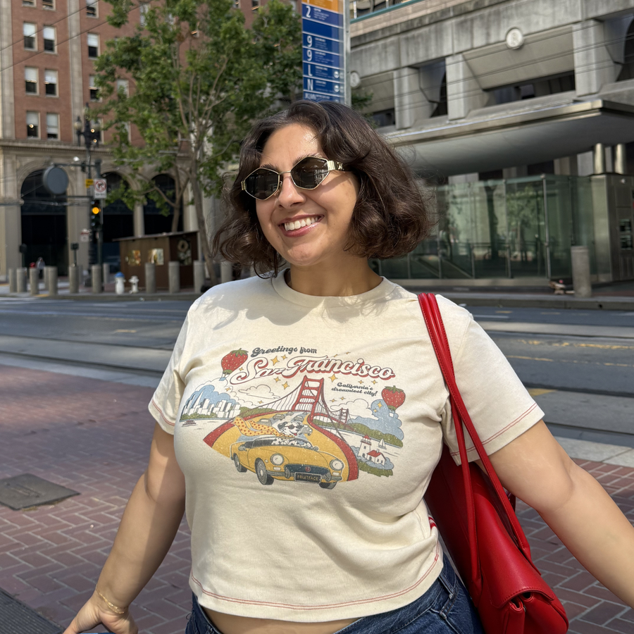Model Jackie smiling on a San Francisco city street wearing the off-white Fruit Face Greetings from SF Baby Tee styled with a red shoulder bag