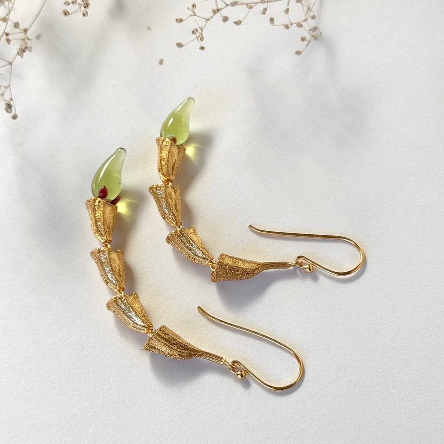 Top-down flat lay view of the Fyoli Fyoli MIRCHI 22K gold-plated chili pod drop earrings with green mouth-blown glass tips on a white background framed by baby's breath flowers.