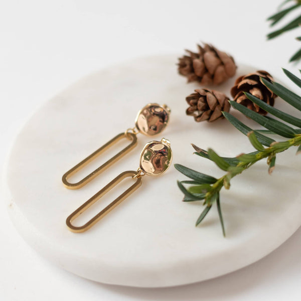 Sela Designs Gold Cleo Earrings featuring textured hammered coin studs with dangling open oval drop pendants on a white marble display.