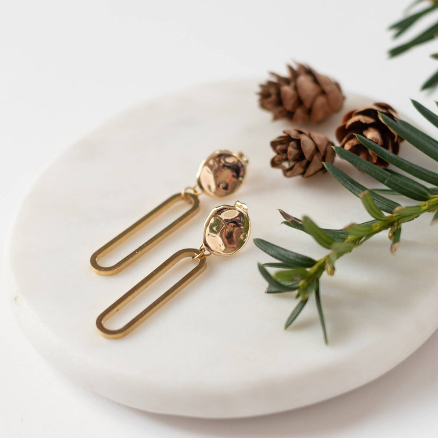 Sela Designs Gold Cleo Earrings featuring textured hammered coin studs with dangling open oval drop pendants on a white marble display.