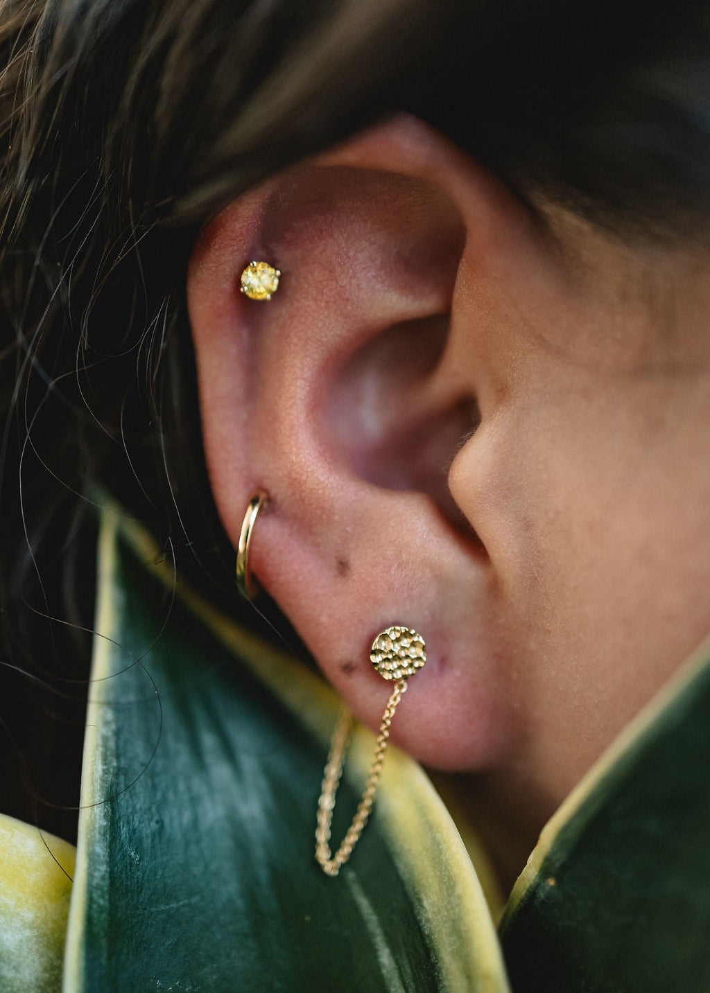 The chain hoop layered on a model's ear alongside a simple huggie hoop and a yellow diamond stud. Leaves are visible in the foreground