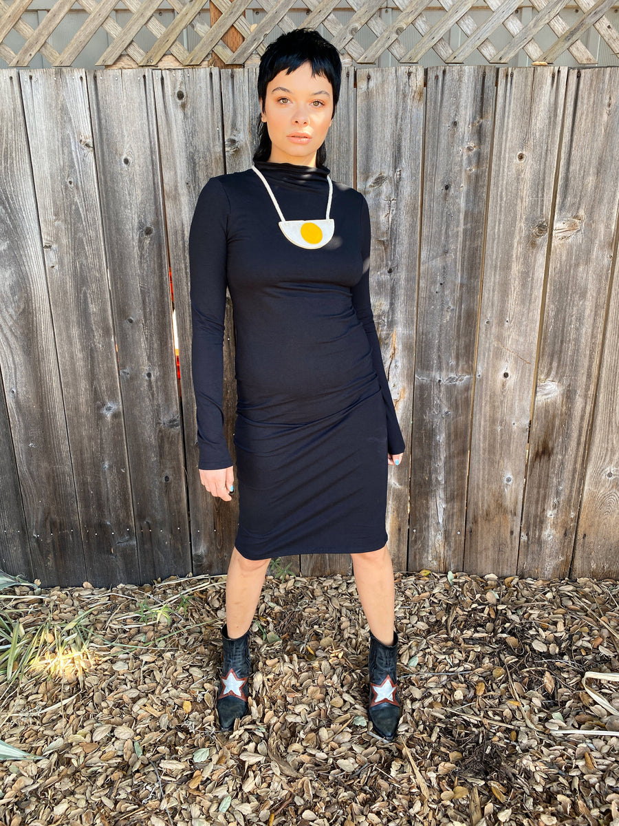 Full-length studio/outdoor portrait of a model posing in the black mock-neck Demi Dress in front of a wooden fence.