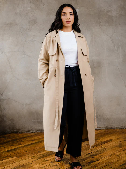 Model wearing the long beige ABLE Kimberly Trench Coat in Brown Sugar over a white top and dark trousers.