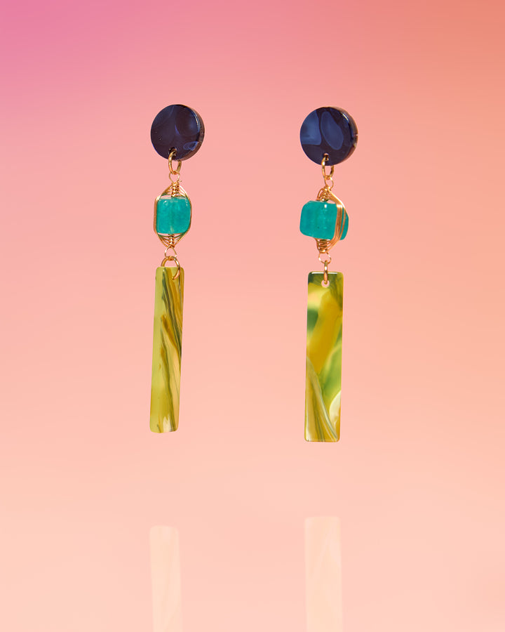 Tribe and Temple Lupe Jade Tropical Dangles featuring a pair of handmade drop earrings with a dark navy blue marbled resin circular post, a wire-wrapped teal jade stone bead, and a long, rectangular lime green and yellow swirled tropical resin bar drop against a soft pink background.