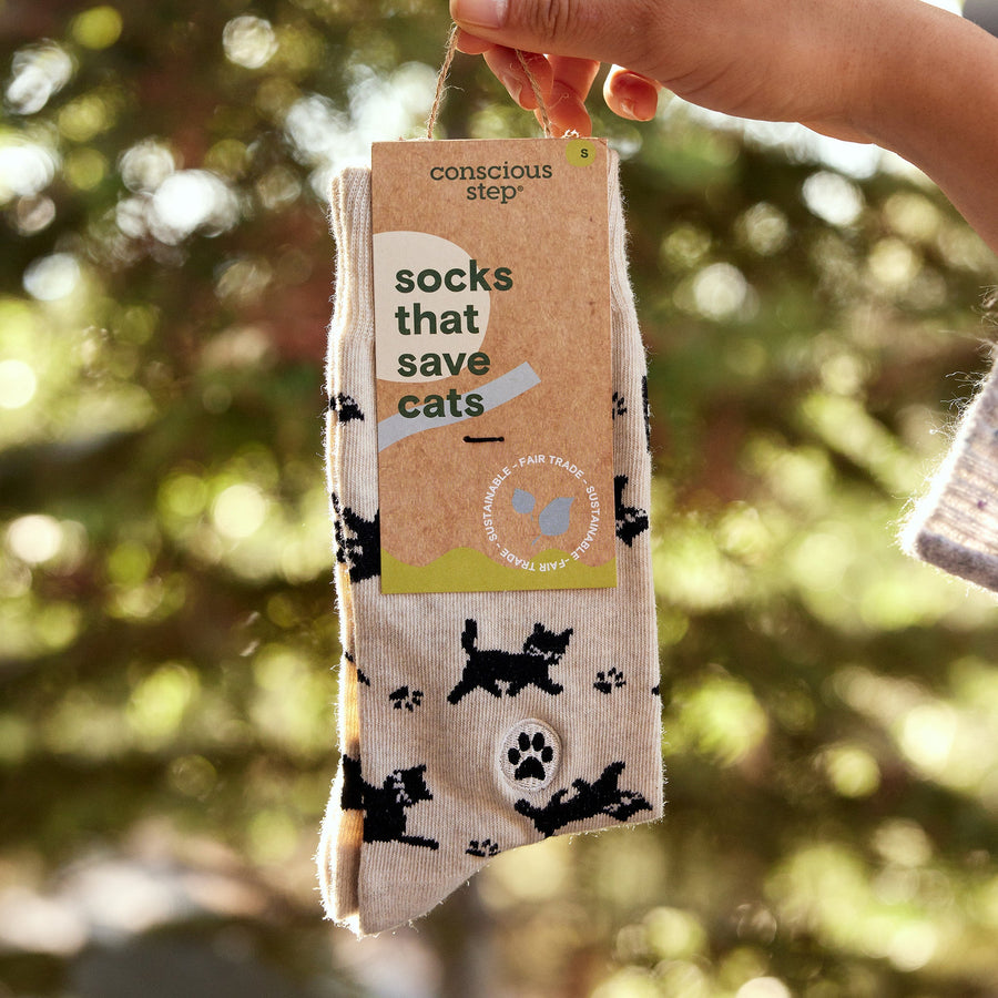 Hand holding a packaged pair of Conscious Step Socks That Save Cats with Fairtrade and organic certification tags against a natural outdoor background.