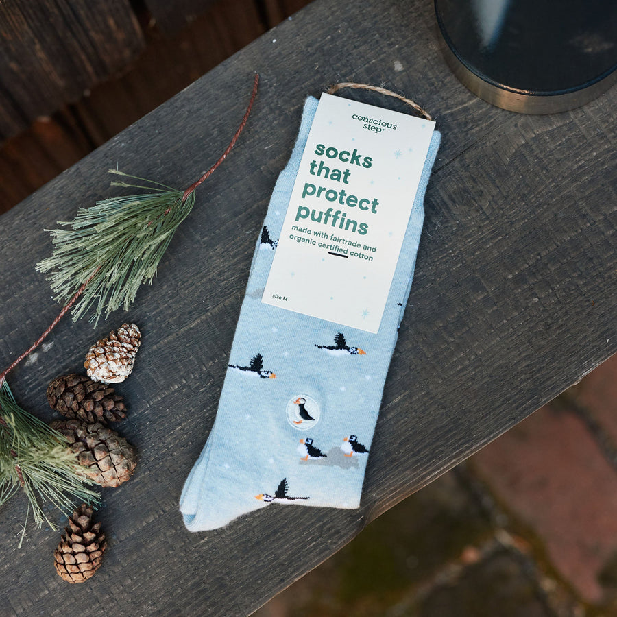 Packaged pair of Conscious Step Socks That Protect Puffins resting on a rustic wooden surface next to pine cones and evergreen sprigs.