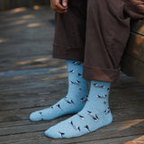 Man sitting on a wooden armchair on a porch wearing a denim jacket, brown trousers, and baby blue Conscious Step puffin socks.